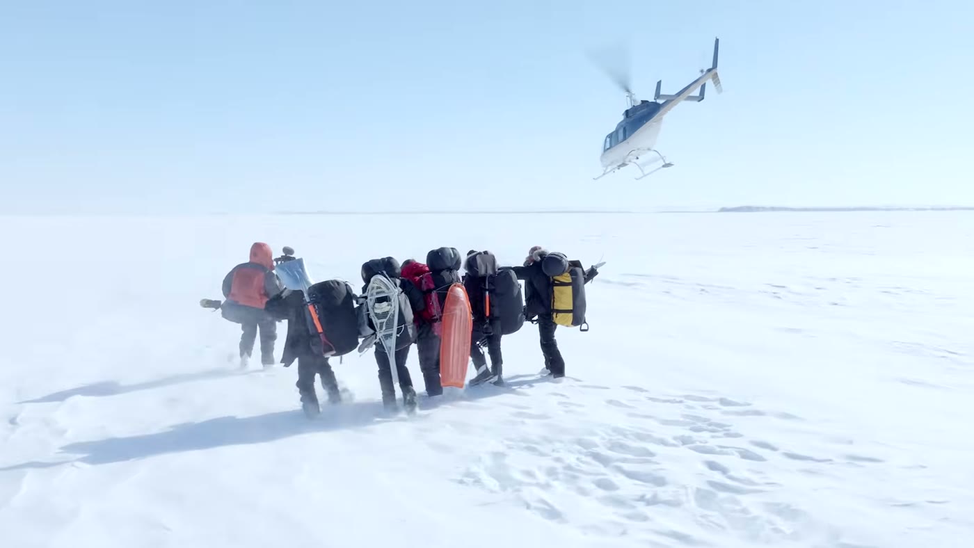 Aerial frame from the analyzed video: a team in cold-weather gear trudges across deep snow as a helicopter lifts off behind them