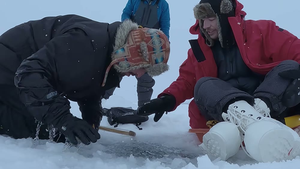 Handheld shot of two people at an ice-fishing hole, captured mid-motion