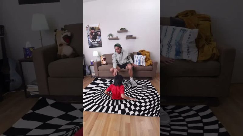 Vertical Short of Zach King reaching toward a child sitting on an optical-illusion checkerboard rug