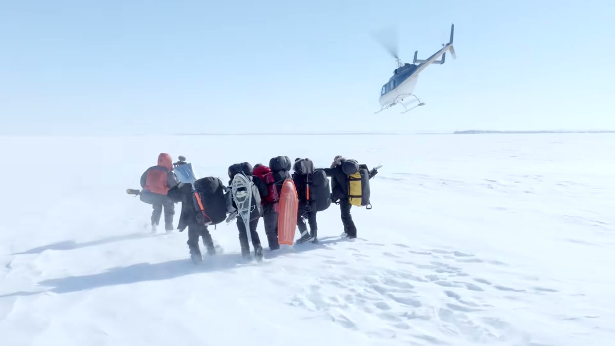 A team in cold-weather gear walks across deep snow as a helicopter takes off behind them