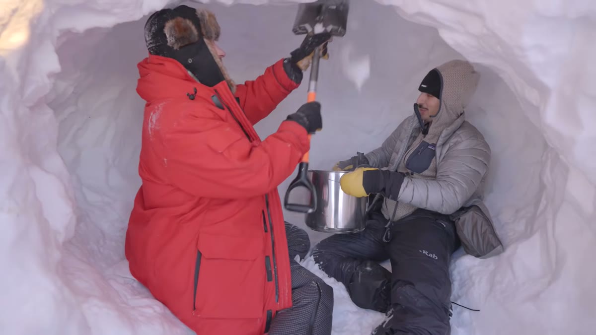 Two people inside a snow shelter, one in a red parka digging with a shovel